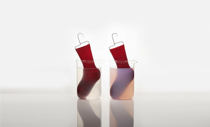 Straight-on shot of two red socks in beakers of water. Red sock on the right bleeding dye. Comparing color bleed between Dirty Labs detergent and conventional detergent.