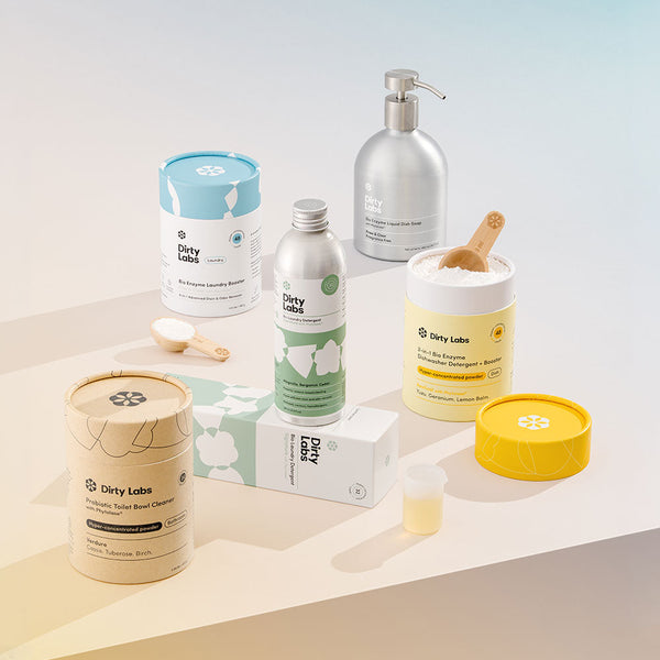 A collection of Dirty Labs cleaning products, including shampoo and laundry booster, displayed on a white surface. The background features a soft pink and green gradient.