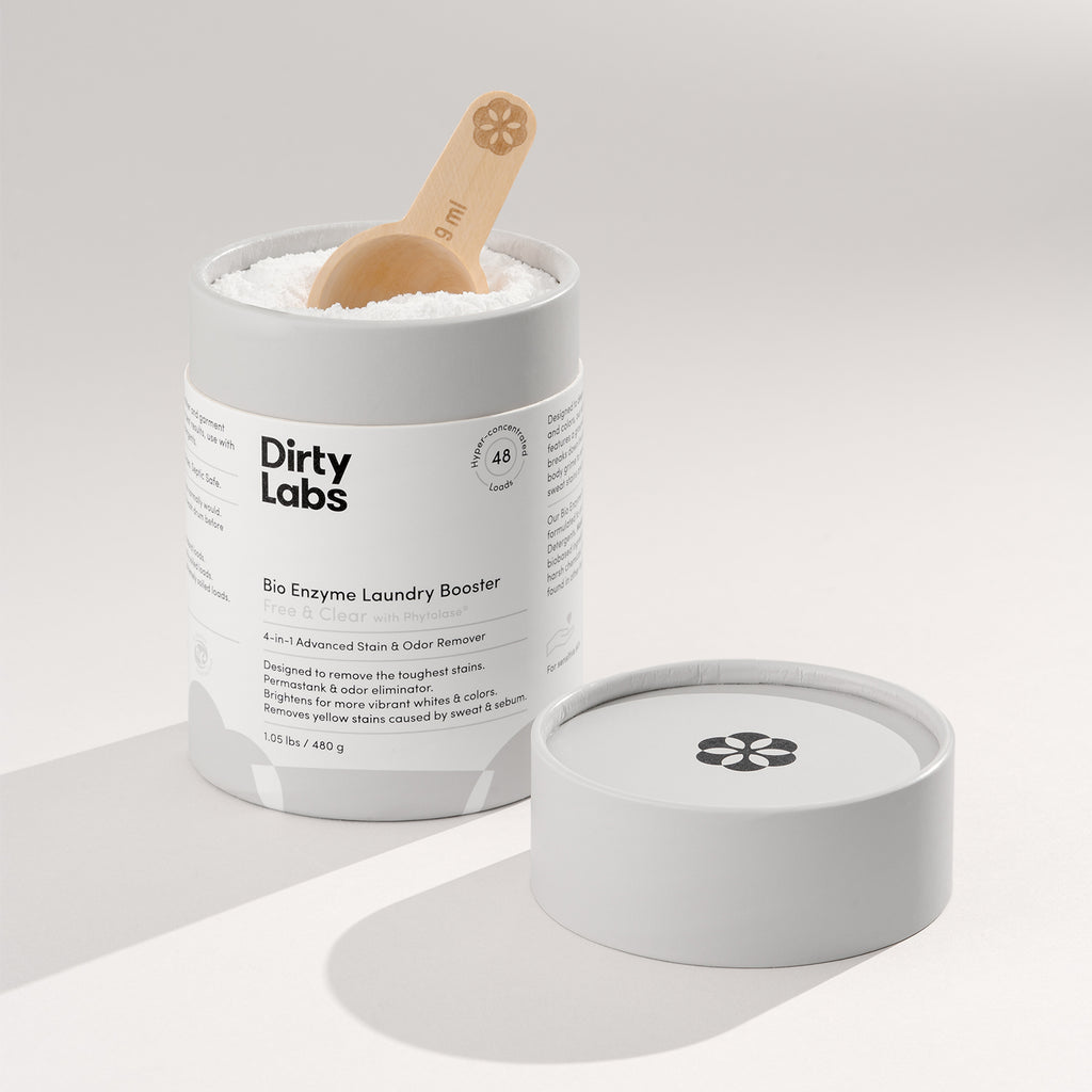 Open white container of Dirty Labs Bio Enzyme Laundry Booster sits on a light surface. A wooden scoop rests in powder, with the lid placed beside. Clean, minimal aesthetic.