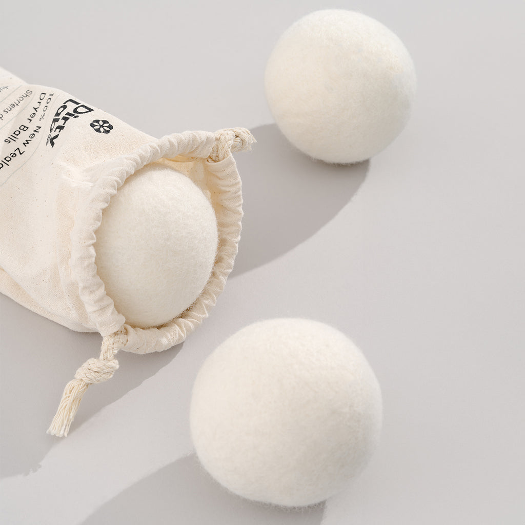 A partially open drawstring bag reveals a white wool ball, with two additional wool dryer balls placed nearby on a plain light surface.