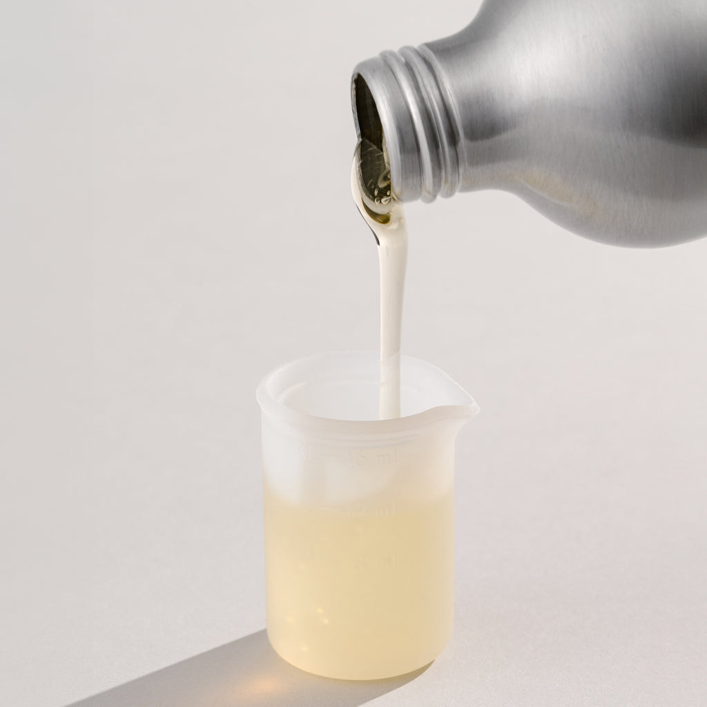 An aluminum bottle pours a thick, pale yellow liquid detergent into a transparent measuring beaker against a neutral grey backdrop.