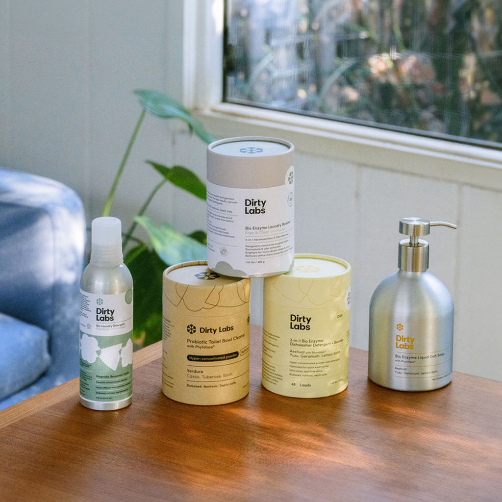5 Dirty Labs detergents resting on a wood table. Far left: signature laundry detergent, Far right: liquid dish soap, middle: toilet bowl cleaner, laundry booster, and automatic dishwasher detergent.