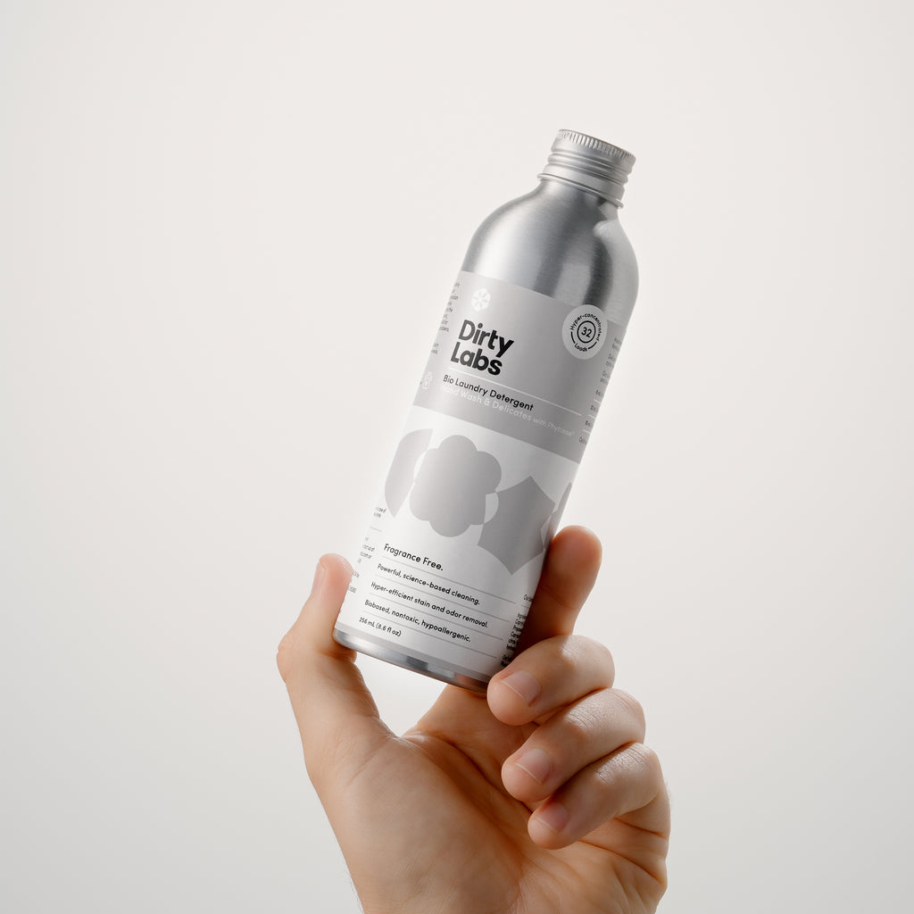 A hand holds a 32-load bottle of Hand Wash & Delicates Bio Laundry Detergent, showcasing the small-size. The bottle has a gray and white label and notes that it's fragrance free. The presentation is displayed against a neutral light grey background.