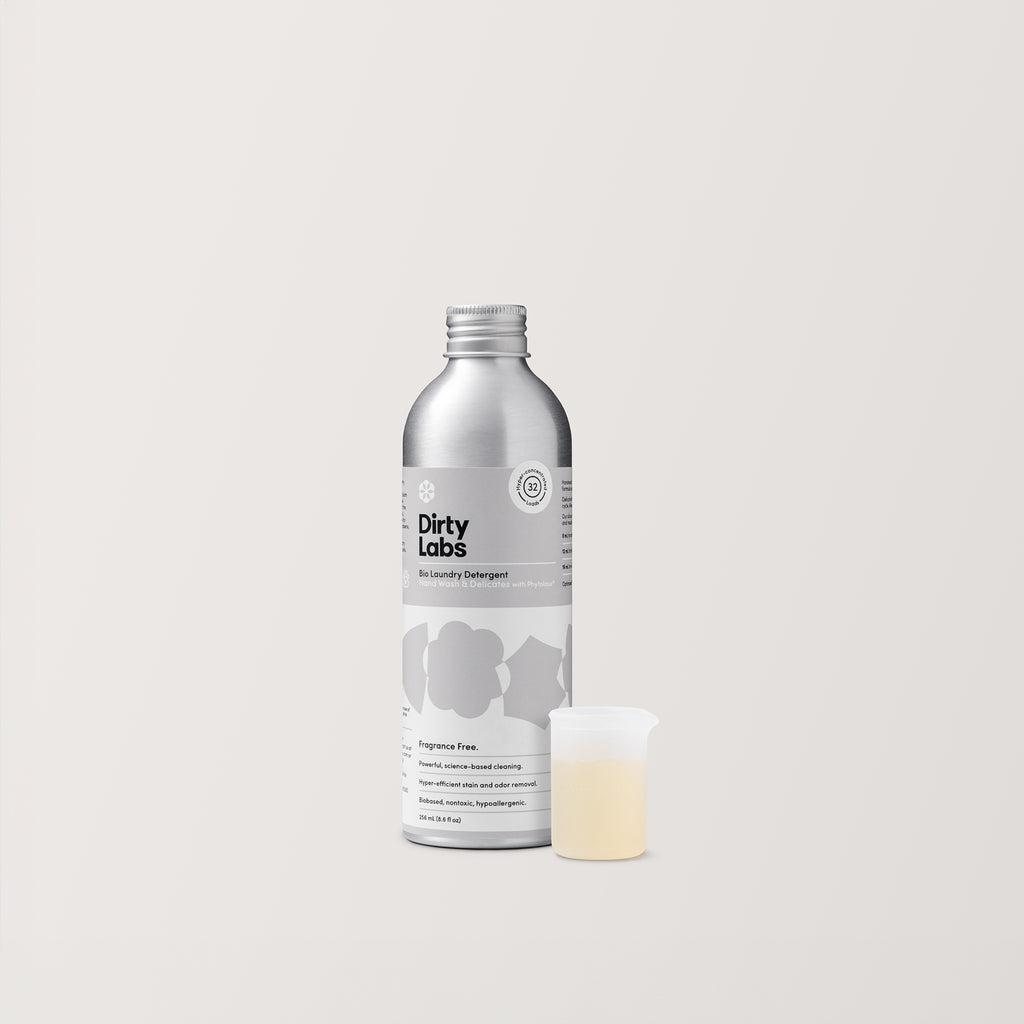 An aluminum 32-load bottle of Dirty Labs Hand Wash & Delicates Bio Laundry Detergent centered against a light grey background. The bottle has a white and gray label and notes that it's fragrance free. Beside the bottle, there is a small silicon measuring beaker filled with yellow liquid.