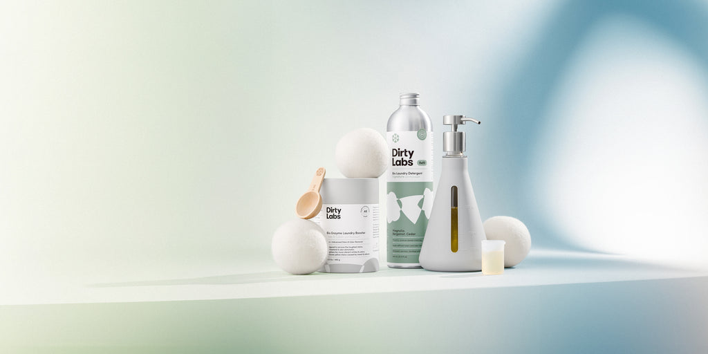 The Sustainable Laundry Starter Set - Signature includes an 80-load bottle of Signature Bio Laundry Detergent, Bio Enzyme Laundry Booster, a reusable flask with a pump, three wool dryer balls, a wooden measuring scoop, and a silicon measuring beaker. The products are showcased against a light grey to blue gradient background.