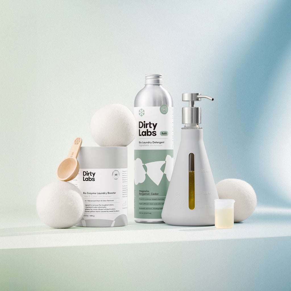 The Sustainable Laundry Starter Set - Signature includes an 80-load bottle of Signature Bio Laundry Detergent, Bio Enzyme Laundry Booster, a reusable flask with a pump, three wool dryer balls, a wooden measuring scoop, and a silicon measuring beaker. The products are showcased against a light grey to blue gradient background.