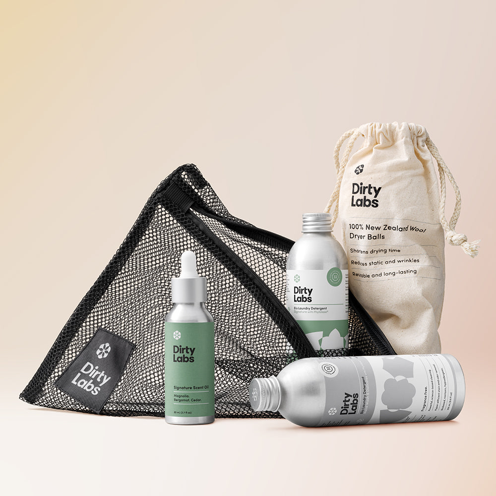 Dirty Labs gift bundle products including 2 detergent bottles and scent oil bottle with a drawstring wool balls bag and black mesh wash bag on an orange gradient background