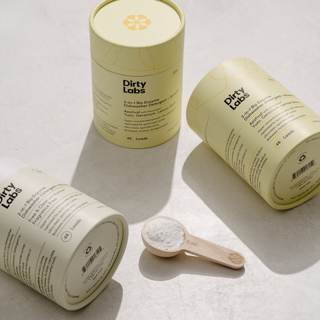 3 Dirty Labs 2-in-1 Bio Enzyme Dishwasher Detergent containers. 2 lay on their sides and one stands upright. There is also a wooden scoop that is full of white powder.