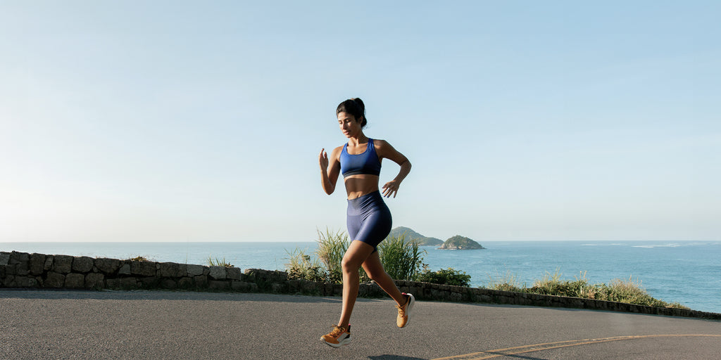 Woman running along a coastal highway. She's wearing blue workout gear and orange shoes. There is blue water and two small islands in the background.