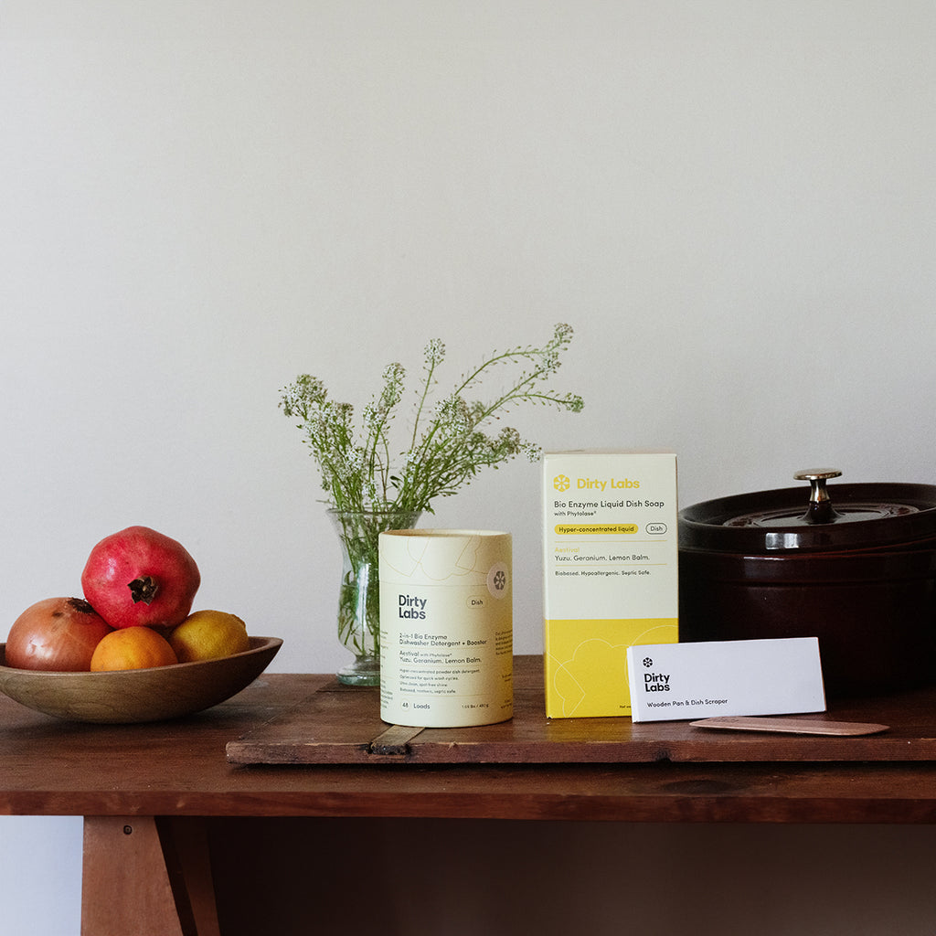 Dirty Labs products on wood table including aestival dishwasher detergent, aestival liquid dish soap yellow box, and wood scraper next to a bowl of apples and green plant.