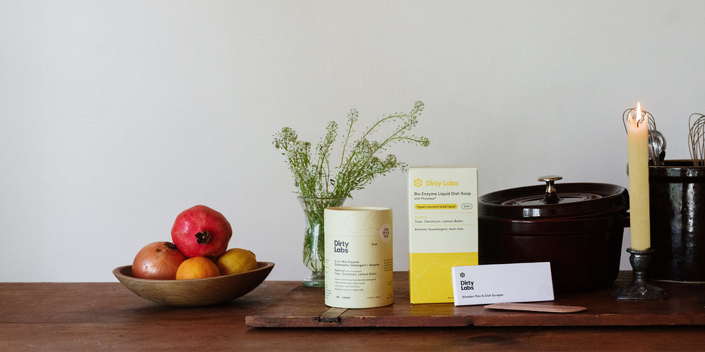 Dirty Labs products on wood table including aestival dishwasher detergent, aestival liquid dish soap yellow box, and wood scraper next to a bowl of apples and green plant.