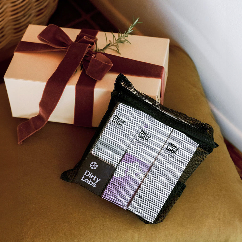 bio laundry holiday bundle 3 pack detergent bundle inside black mesh bag. Bag is resting against brown box with red bow on brown felt chair.