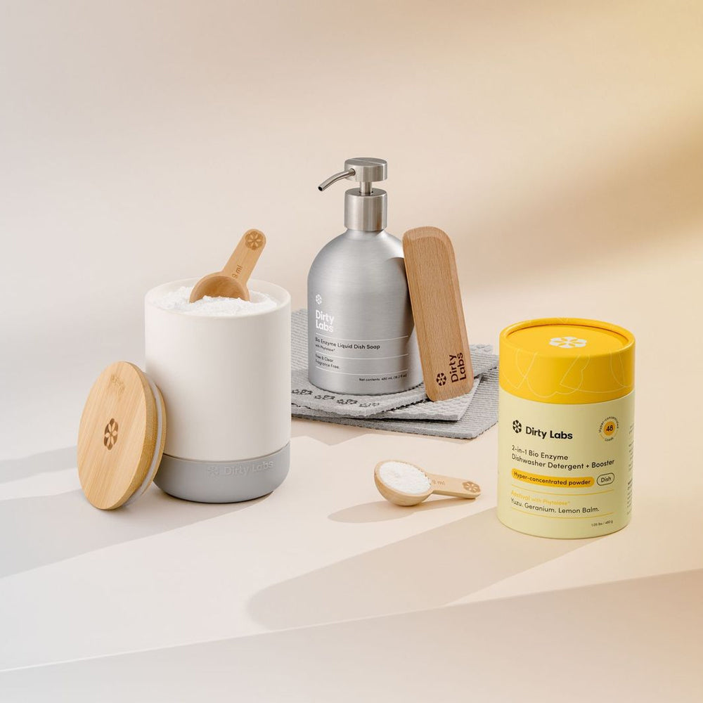 Collection of Dirty Labs' products including a white ceramic canister, silver liquid dish soap bottle with wooden scraper, and yellow dishwasher powder container on a light background.
