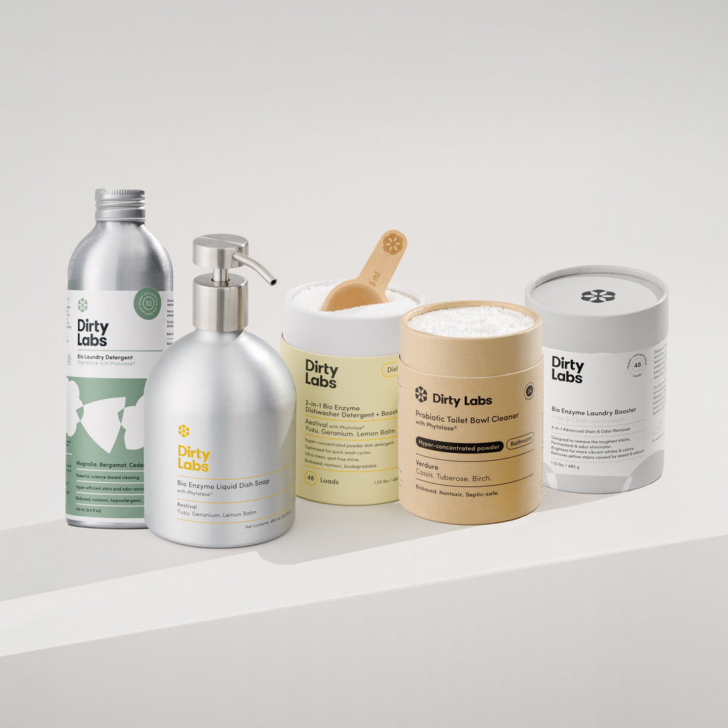 The Essentials Starter Set from Dirty Labs features eco-friendly laundry detergent, probiotic toilet bowl cleaner, dish soap, and more, arranged side by side on a white surface with their labels visible against a light background.