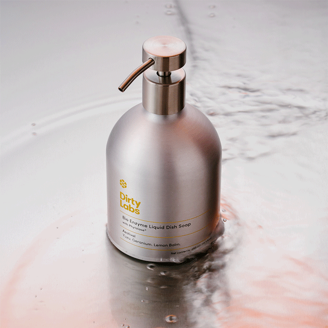 A silver pump bottle of Aestival Bio Enzyme Liquid Dish Soap stands on a wet surface with water ripples. The label highlights its Summer Scent—Geranium, Lemon Balm—and features advanced Phytolase® enzyme technology.