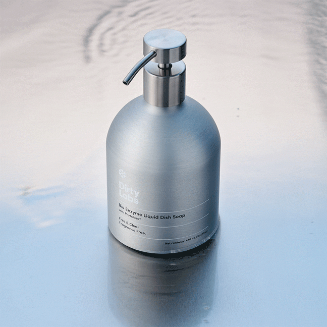 A stainless steel pump bottle labeled Free & Clear Bio Enzyme Liquid Dish Soap, featuring biodegradable detergent with Phytolase® enzyme technology, stands on a reflective, slightly rippled surface under soft lighting.