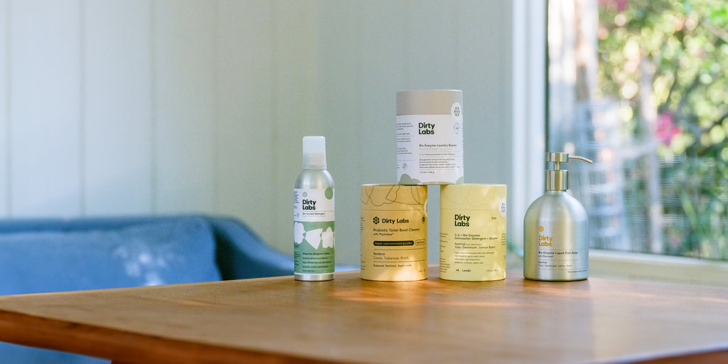 Five Dirty Labs cleaning products, including liquid detergent, powders, and a pump dispenser, arranged on a wooden table in a sunlit room with a blue couch and window in the background.