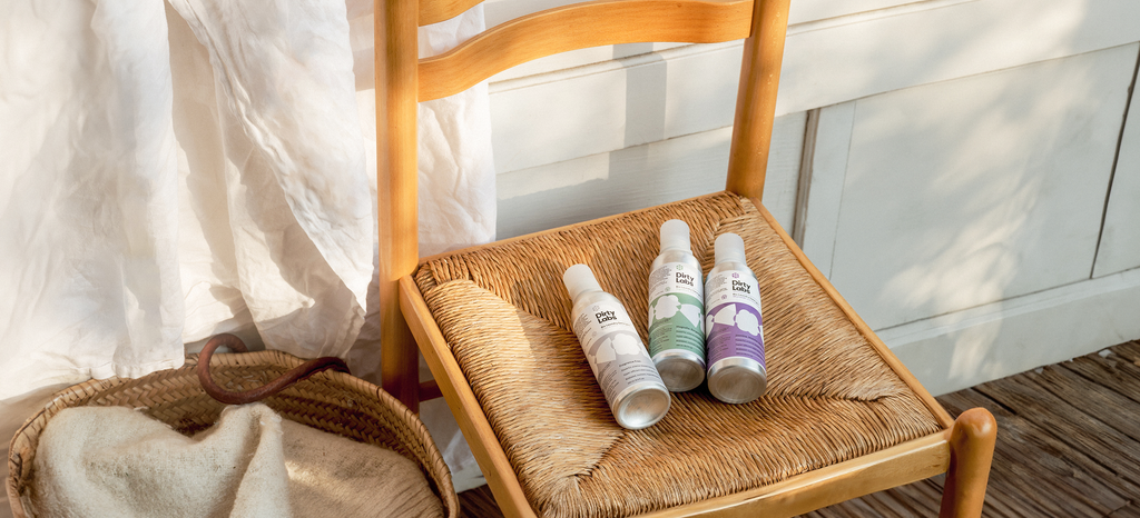 Close up of 3 Dirty Labs 32-load detergent bottles on a wood rattan chair with a laundry basket to the left against a wall with sunlight.