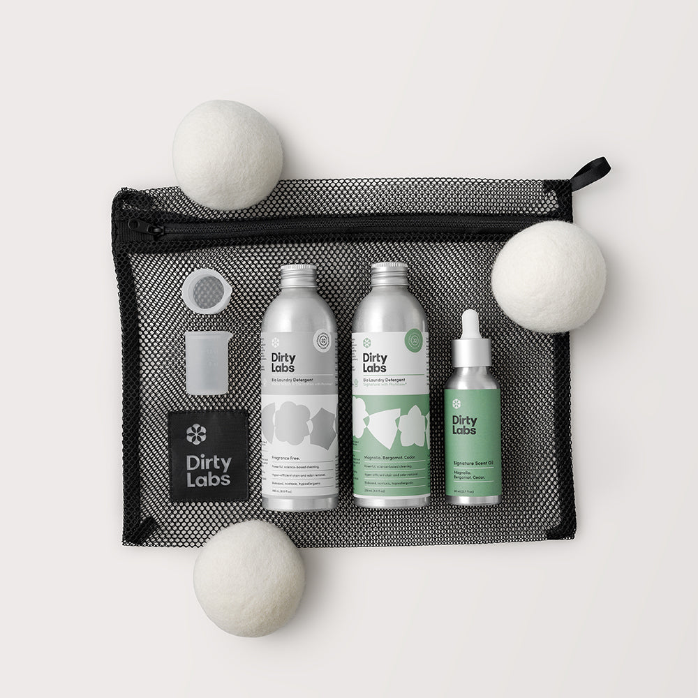 Set of Dirty Labs cleaning products including bottle, scent oil, and wool balls in a mesh bag on a light grey background background