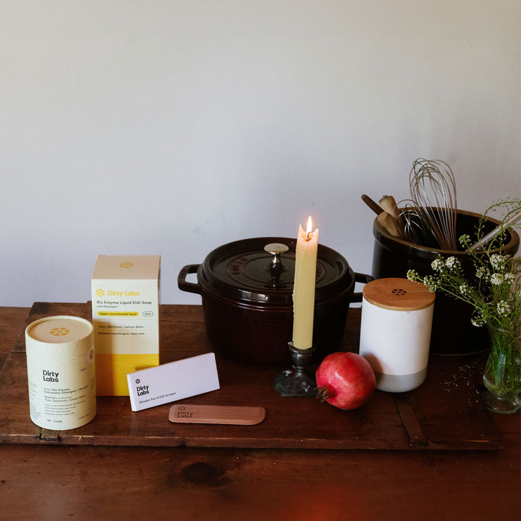 Dirty Labs aestival automatic dishwasher detergent container alongside Dirty Labs liquid dish soap yellow box and wood scraper on cherry wood cutting board and table with candle and apple.