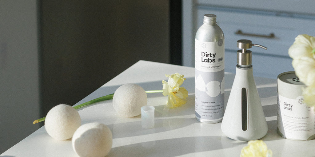 Dirty Labs 80 load detergent with reusable pump, oxygen booster, and wool dryer balls next to yellow flowers on white table with sunlight shining.