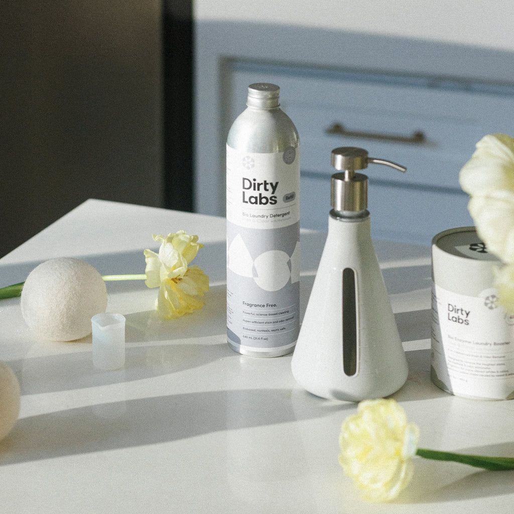 Dirty Labs 80 load detergent with reusable pump, oxygen booster, and wool dryer balls next to yellow flowers on white table with sunlight shining.