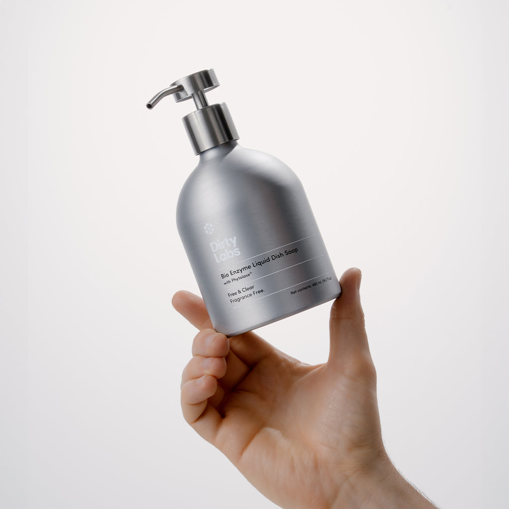 A hand holds a silver pump bottle labeled Free & Clear Bio Enzyme Liquid Dish Soap, showcasing a sleek, minimalist design and ultraconcentrated formula against a plain light background.