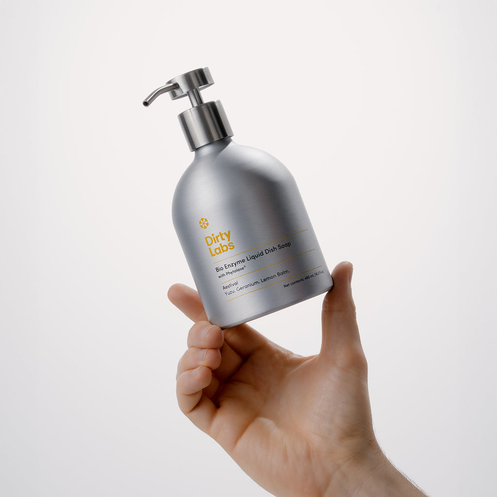 A hand holds a silver pump bottle of Aestival Bio Enzyme Liquid Dish Soap against a plain, light background. The label mentions the scent: Wild Lavender, Lemon Zest.