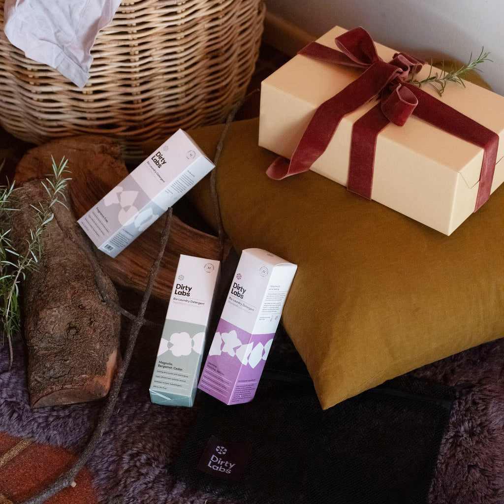Cardboard box wrapped with red ribbon on yellow pillow with Dirty Labs 32 load laundry detergent boxes resting against the pillow with wood log.