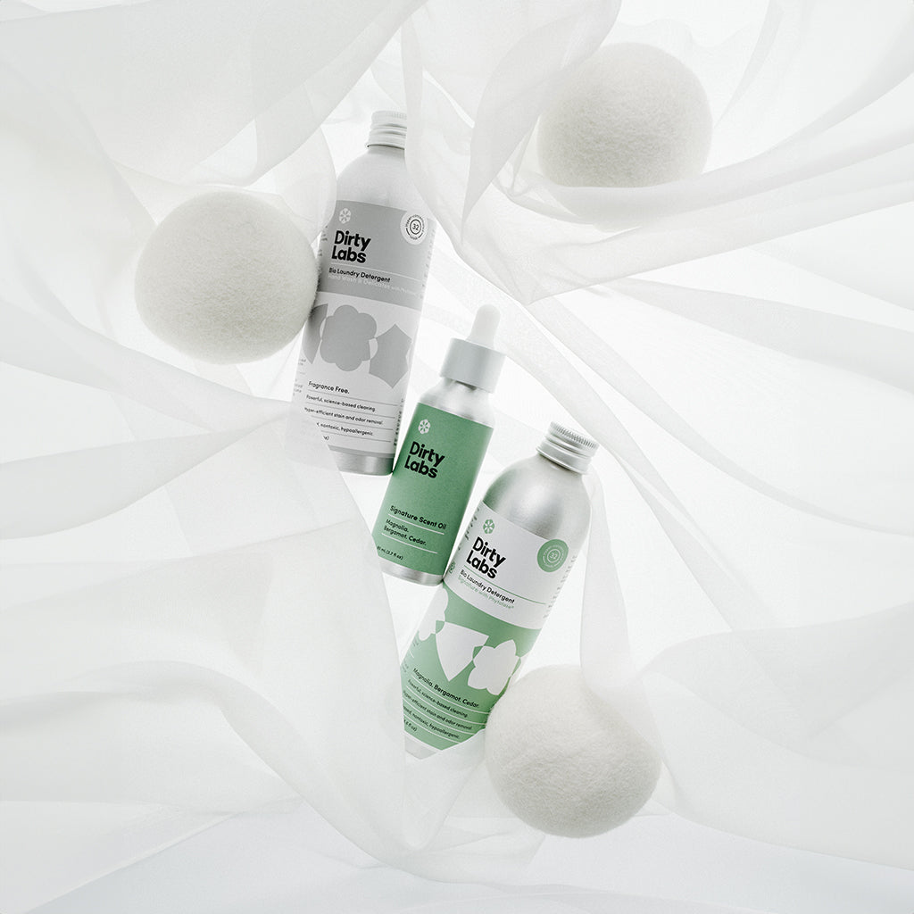 Dirty Labs Hand wash 32 load next to Signature scent oil and 32 load Signature detergent with Wool Dryer Balls on a light soft texture background