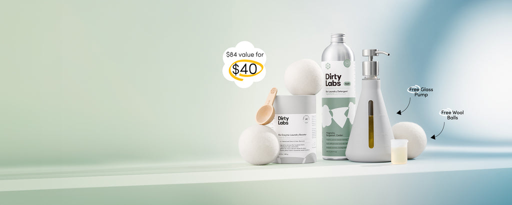 Dirty Labs 80 load signature detergent bottle with green label standing next to Booster powder container and Glass pump and wool balls against a gradient green and blue backdrop. Text on image highlights $84 value for $40.