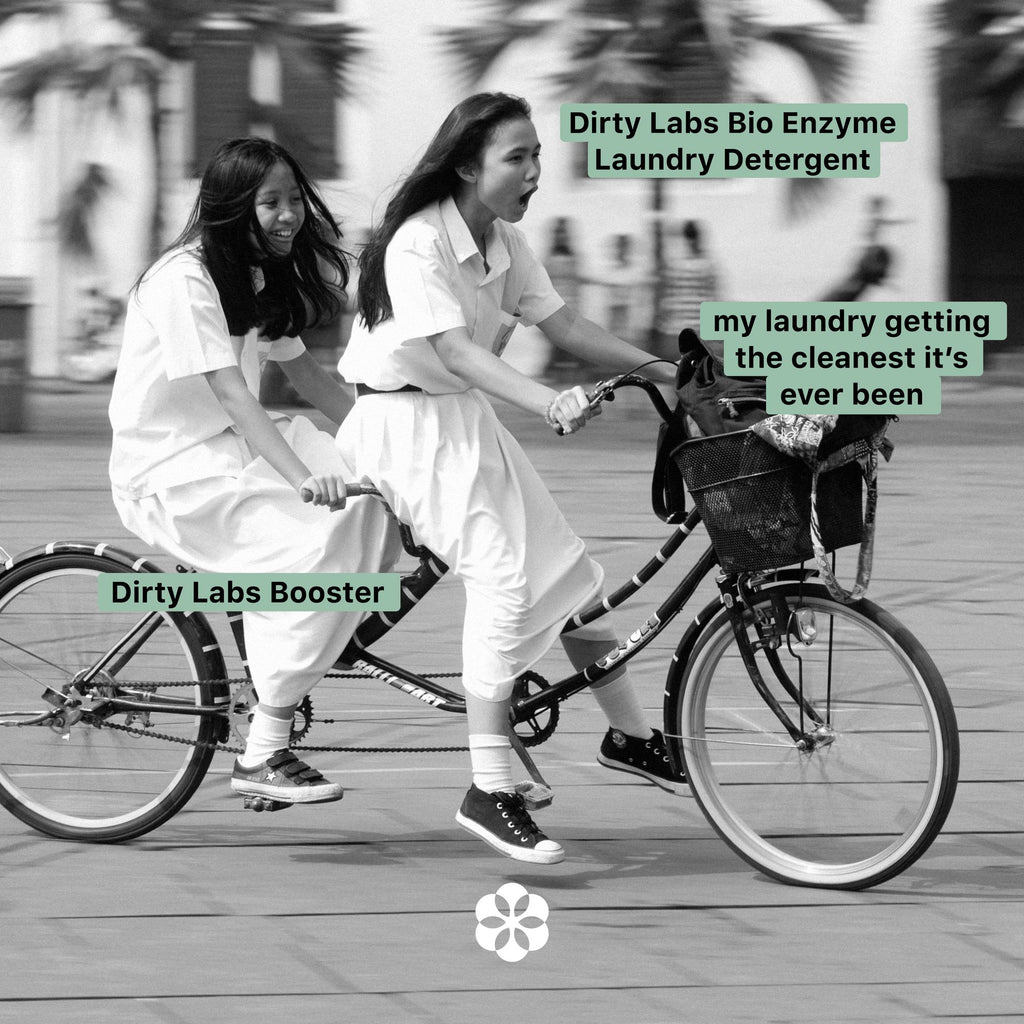 Black and white image of two young girls riding a tandem bike. The girl in front is labeled 'Dirty Labs Bio Enzyme Laundry Detergent' while the girl in back is labeled 'Dirty Labs Booster'. The bike is labeled 'my laundry getting the cleanest it's ever been'.