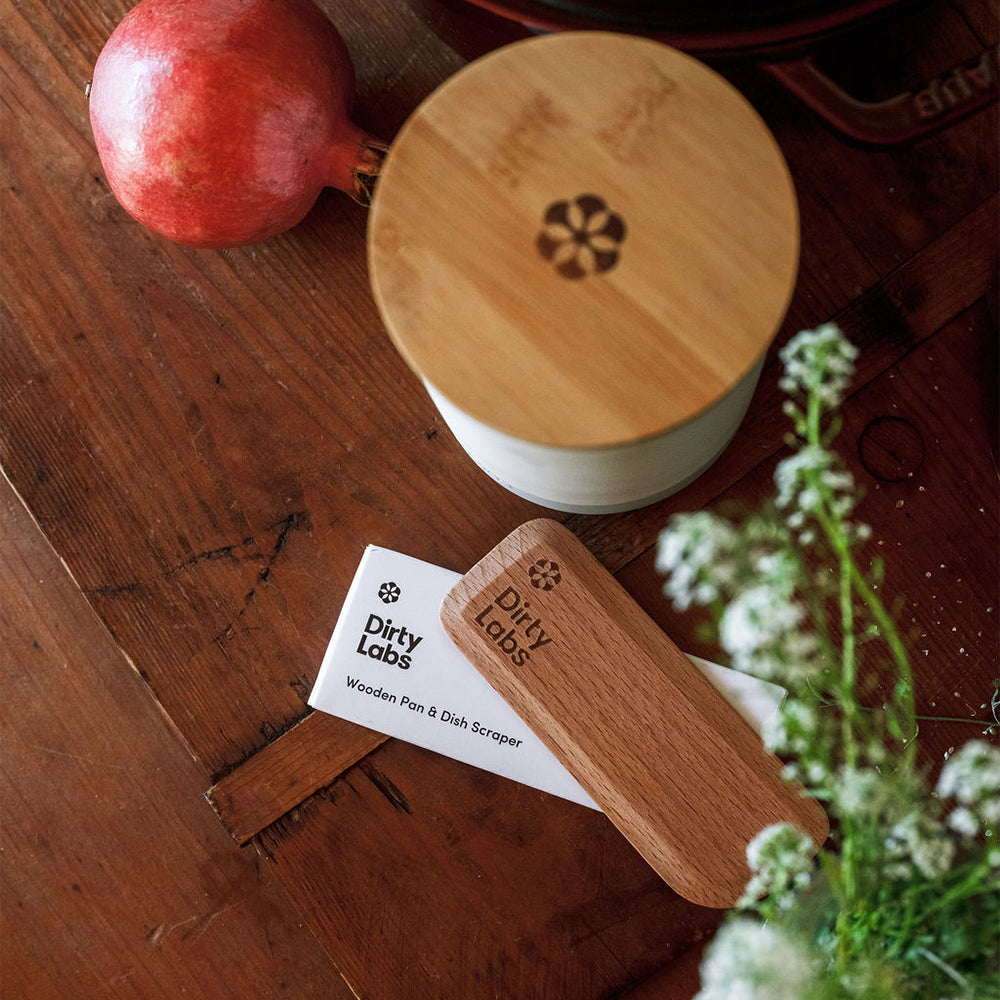 Wooden pan, dish scrubber, and card on a wooden surface with a pomegranate and flowers.