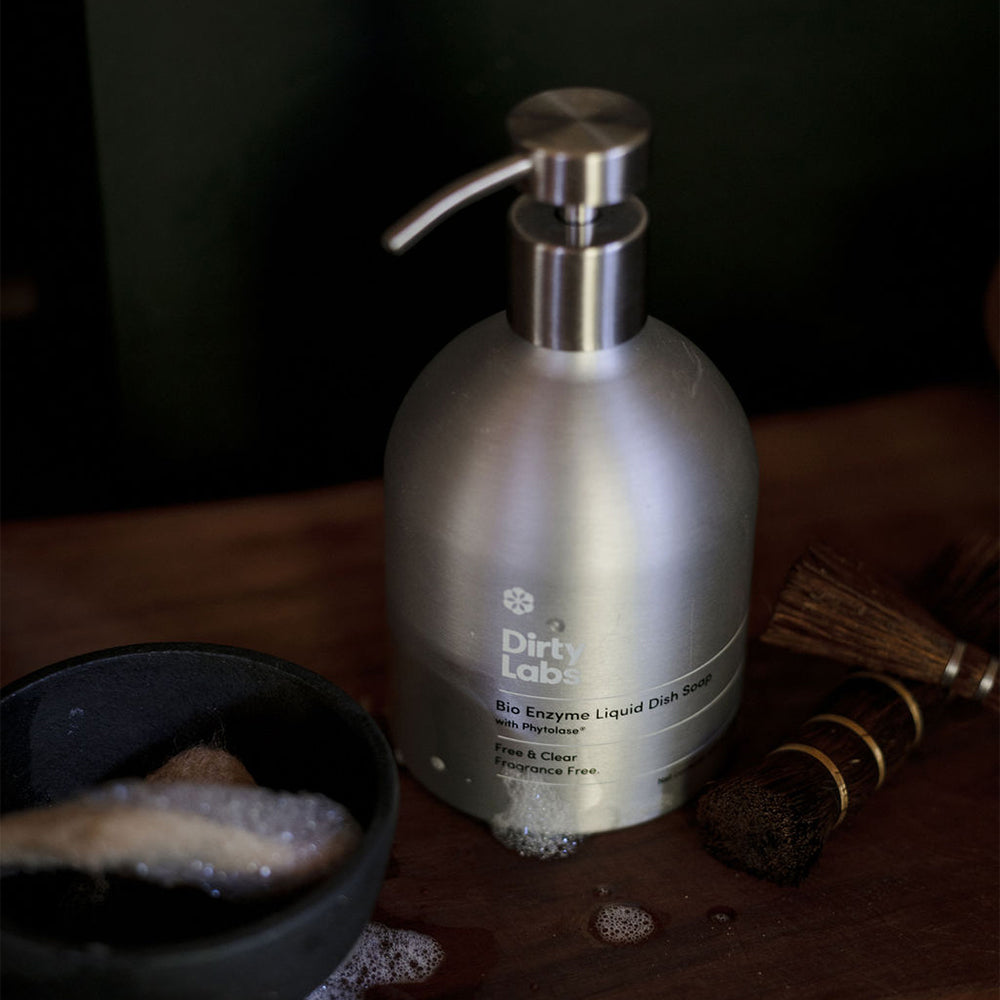 Silver bottle labeled 'Dirty Labs' on a wooden surface with cleaning tools and dish soap bubbles