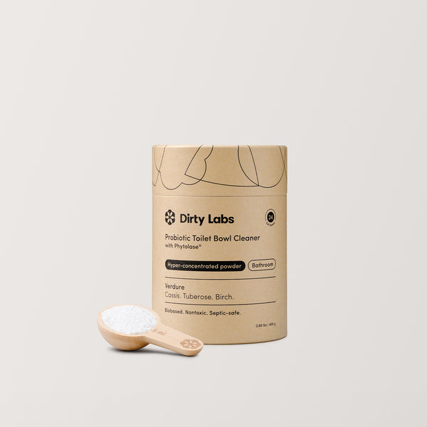 A cylindrical container of Probiotic Toilet Bowl Cleaner with Phytolase™ is set against a light background, next to a wooden scoop filled with cleaning powder. The brown packaging is decorated with black text and illustrations, highlighting its nontoxic formula.