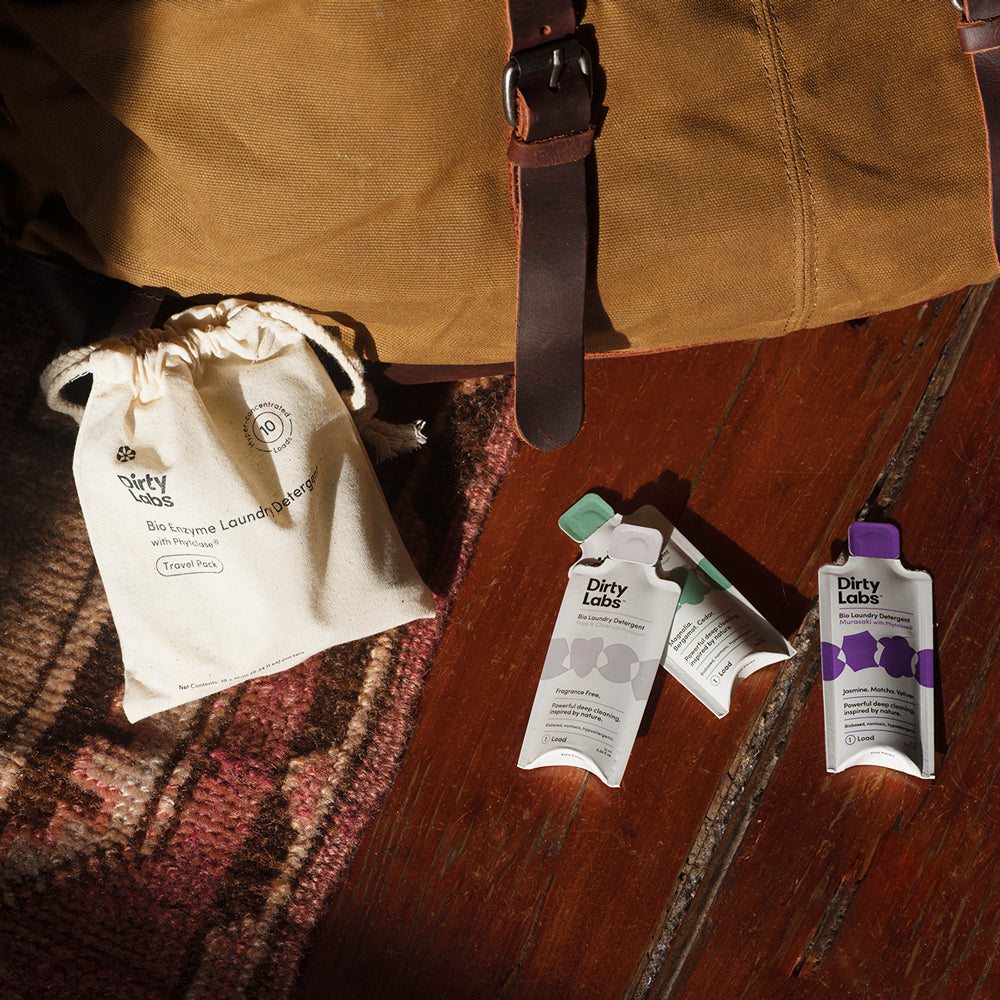 Dirty Labs laundry detergent single dose packets on a wooden surface with rug and travel bag luggage.